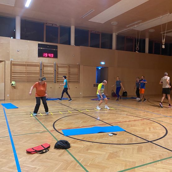 Eine Gruppe von Menschen trainiert in einem Fitnessstudio, wobei eine Person auf einem Stepper steht und andere herumstehen. Eine Digitaluhr zeigt die Zeit 19:49 an.