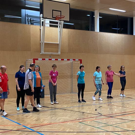 Eine Gruppe von Frauen und einem Mann steht in einer Turnhalle, wahrscheinlich in Vorbereitung auf ein Training. Sie tragen Sportbekleidung und Turnschuhe. Im Hintergrund sind ein Basketballkorb und ein Tor zu sehen.