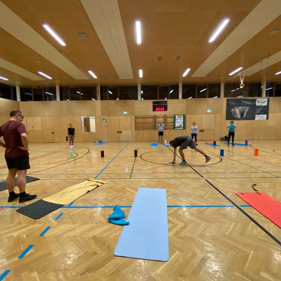 Eine Gruppe von Menschen trainiert in einer Turnhalle mit Holzböden. Einige machen Yoga, während andere ein Spiel mit kleinen Objekten auf dem Boden spielen.