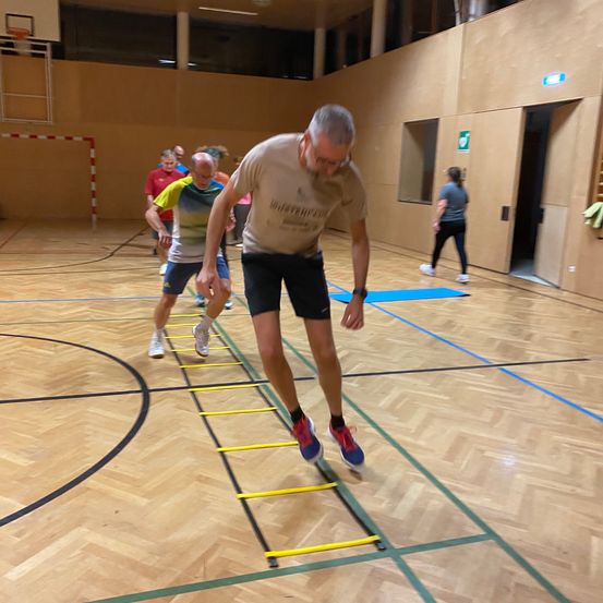 Ein älterer Mann tritt in einer Turnhalle auf eine gelbe Leiter, während andere Personen im Hintergrund trainieren.