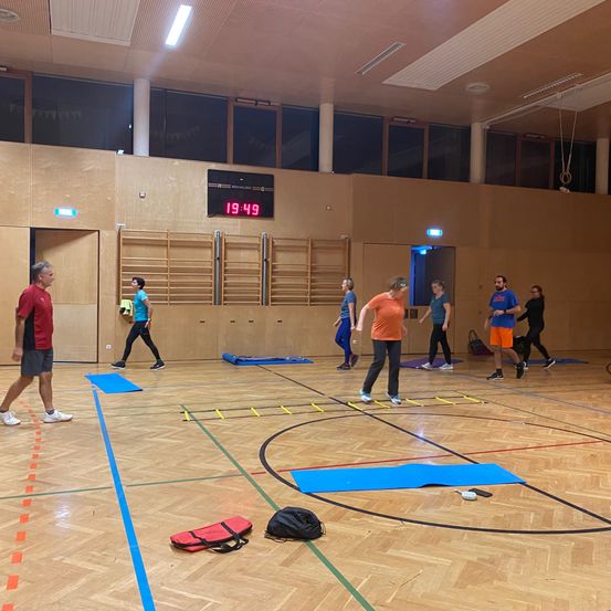Eine Gruppe von Menschen trainiert in einer Turnhalle. Sie laufen auf einem blauen Teppich und benutzen eine Leiter. Es gibt Taschen und Matten auf dem Boden. Die Zeit ist 19:49.