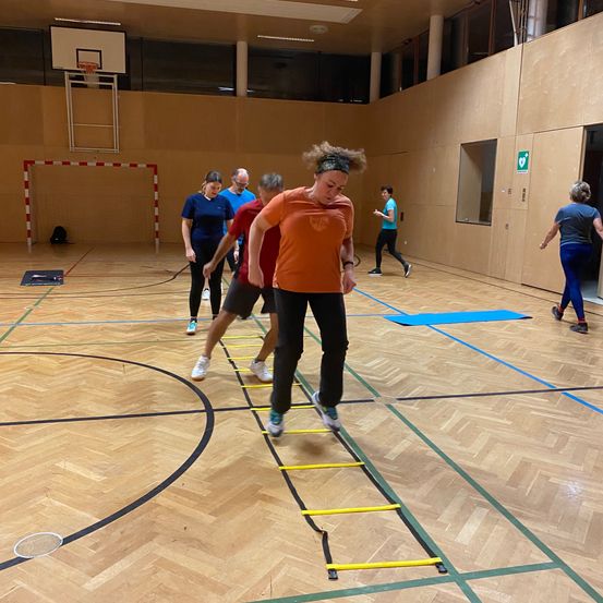 Eine Gruppe von Menschen trainiert in einer Indoor-Sporthalle. Eine Frau steht auf einer gelben Leiter, während andere zusehen. Die Halle hat einen Basketballplatz und einen Korb.