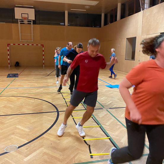 Eine Gruppe von Menschen trainiert in einer Turnhalle, wobei ein Mann auf eine Leiter tritt, während andere zusehen.