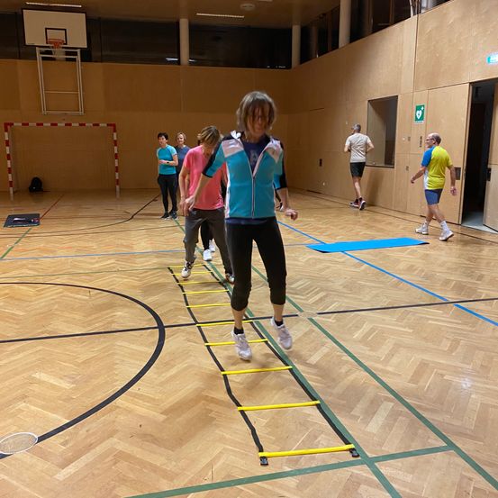 Eine Gruppe von Menschen macht in einer Turnhalle auf einem Holzboden Beweglichkeitsübungen, wobei eine Frau auf eine Leiter tritt.