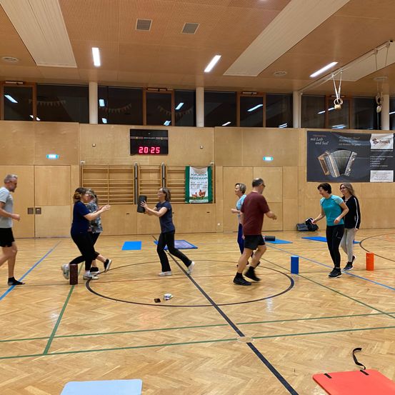 Eine Gruppe von Menschen spielt ein Spiel auf einem Hallenplatz. Sie tragen Turnschuhe und einige haben Armbänder. Eine Anzeigetafel ist an der Wand montiert.