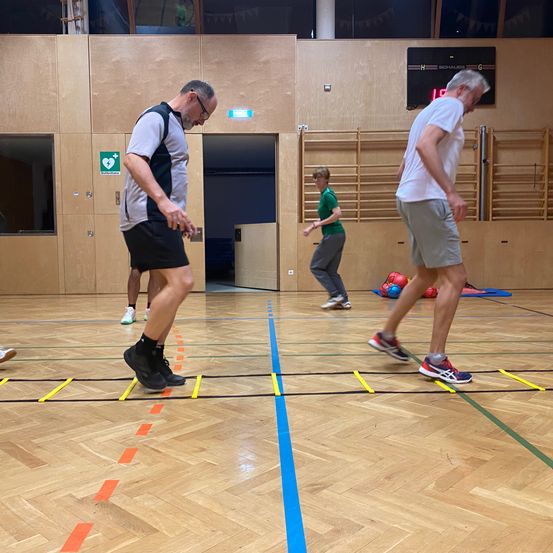 Drei Personen üben auf einem Holzboden mit farbigen Linien Koordination. Ein Mann mit Brille und Turnschuhen tritt auf eine gelbe Linie.