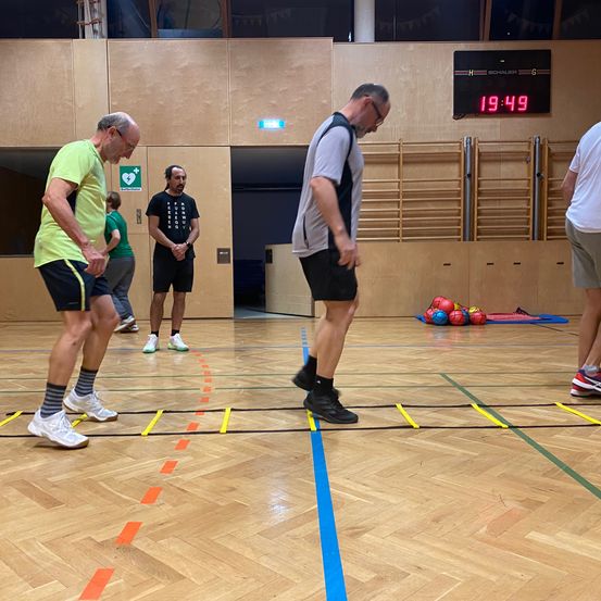Zwei Männer machen Beweglichkeitsübungen auf einem Sporthallenboden, der mit farbigen Linien markiert ist. Eine digitale Uhr zeigt die Zeit an.