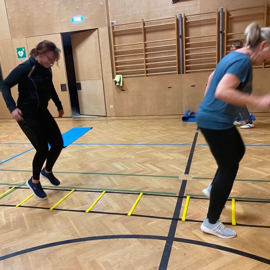 Zwei Frauen führen Geschicklichkeitsübungen auf einem Holzboden durch, der mit farbigen Linien und einer gelben Leiter markiert ist. Eine Frau trägt ein schwarzes Outfit, die andere ein blaues.