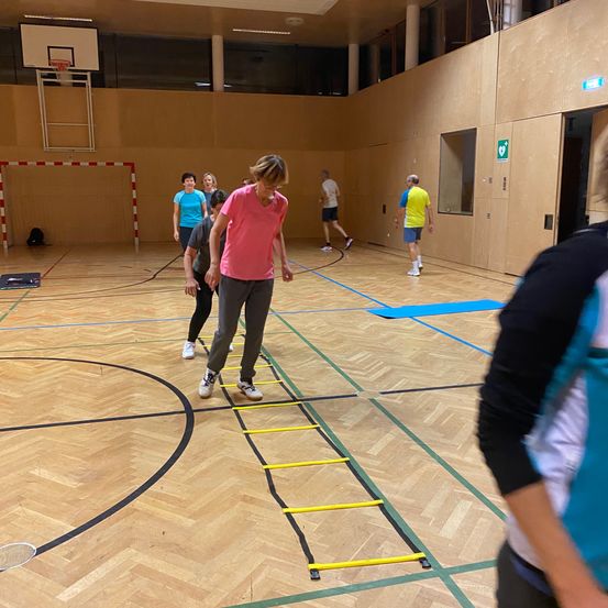 Eine Gruppe von Menschen trainiert in einer Turnhalle. Eine Person klettert eine gelbe Leiter, während andere herumstehen. Die Turnhalle hat einen Holzboden, einen Basketballkorb und eine Tür.