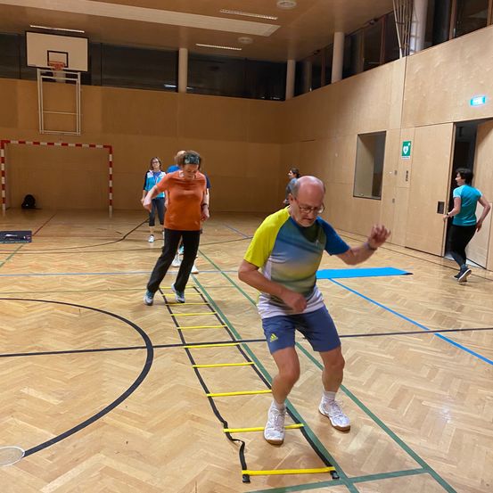 Eine Gruppe von Menschen trainiert in einer Turnhalle. Ein älterer Mann klettert eine Leiter, während andere in der Nähe gehen. Die Turnhalle hat Holzböden und Wände.