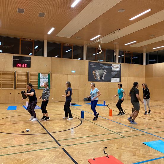 Eine Gruppe von Frauen trainiert in einer Turnhalle mit Holzböden, hält Hanteln und Matten. Eine Digitaluhr zeigt die Zeit an.