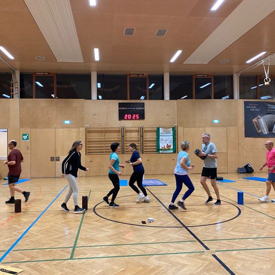 Eine Gruppe von Menschen trainiert in einer Turnhalle, trägt Turnschuhe und hält Hanteln. Eine Digitaluhr zeigt die Zeit 20:25 an. Die Turnhalle hat Holzböden und Wände.