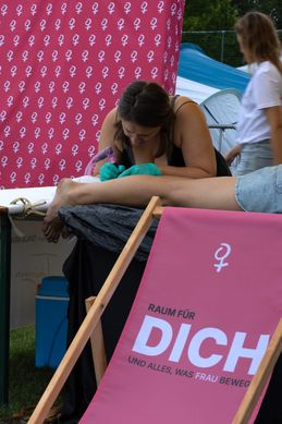 Eine Frau tätowiert ein Bein auf einem Event im Freien. Die Person liegt auf einem Tisch mit einem pinken Schild, das 'Raum für Dich' besagt.