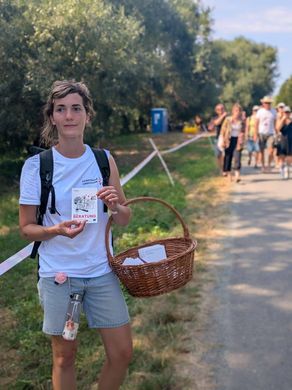 Eine Frau in einem weißen T-Shirt hält einen Korb und eine Karte mit der Aufschrift 'BERATUNG', während sie auf einem Pfad steht, hinter dem Menschen gehen.