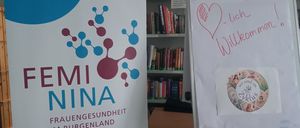 Ein Banner für Femi Nina steht neben einem Whiteboard mit einem Herz und der Aufschrift 'Herzlich willkommen'. Hinter dem Whiteboard befindet sich ein Bücherregal aus Holz.