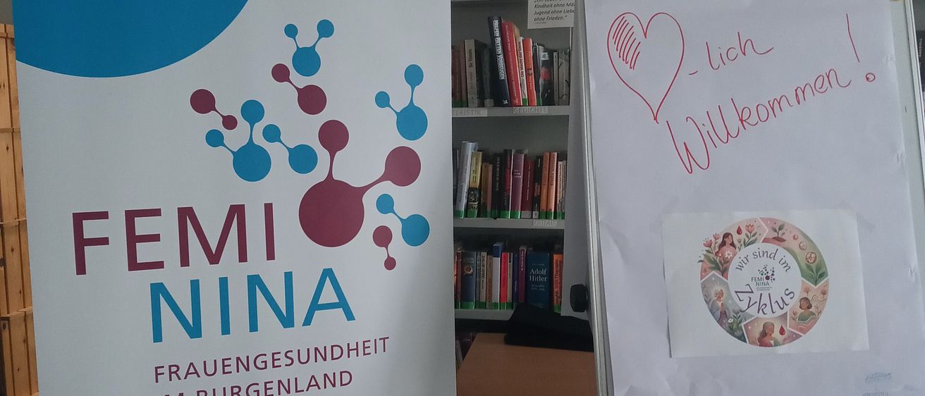 Ein Banner für Femi Nina steht neben einem Whiteboard mit einem Herz und der Aufschrift 'Herzlich willkommen'. Hinter dem Whiteboard befindet sich ein Bücherregal aus Holz.