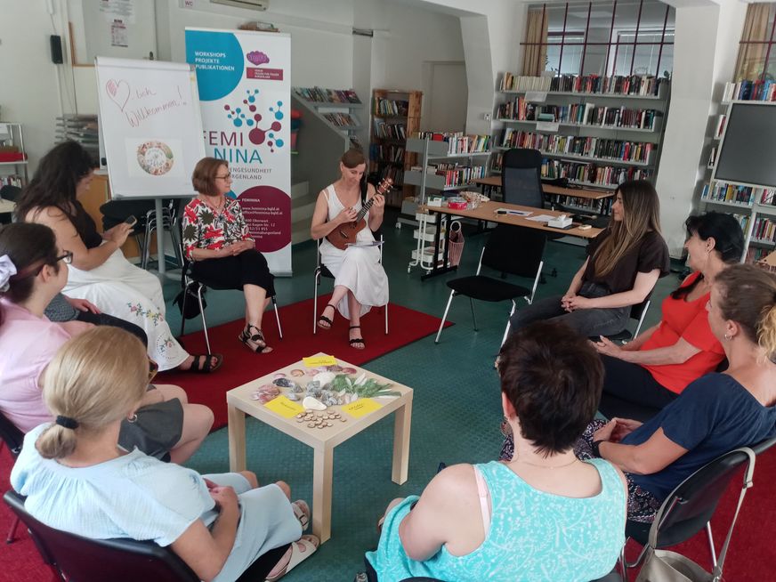Eine Frau in einer Bibliothek spielt Ukulele, während eine Gruppe von Menschen zuhört. Einige sitzen auf Stühlen um einen Tisch, andere auf dem Boden. Es gibt eine Tafel und einen roten Teppich.