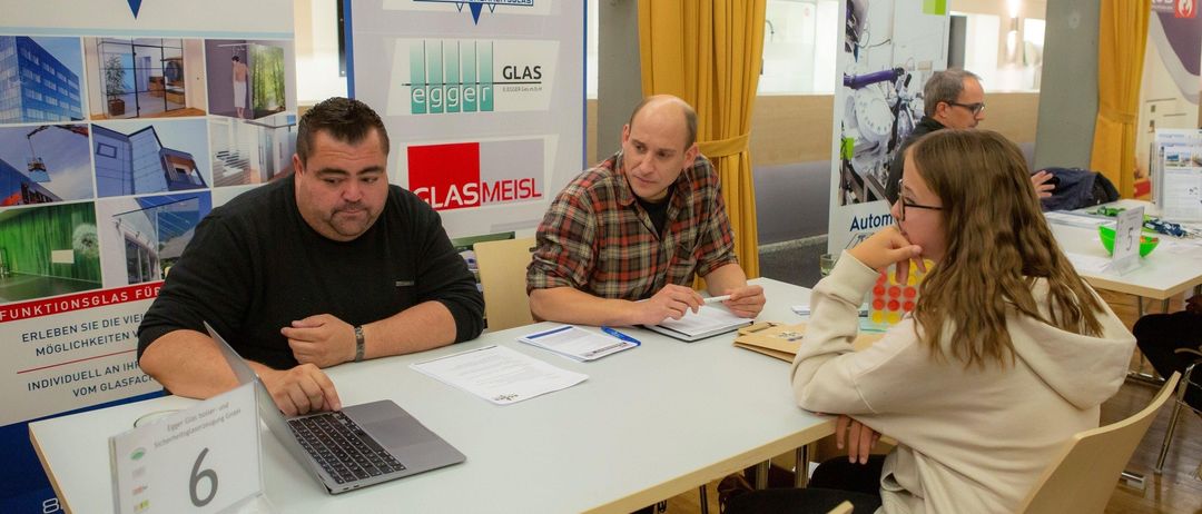 Drei Personen sitzen an einem Tisch mit Laptops und Papieren. Zwei Männer schauen eine Frau an, die eine Brille trägt. Hinter ihnen befinden sich Banner mit Logos.