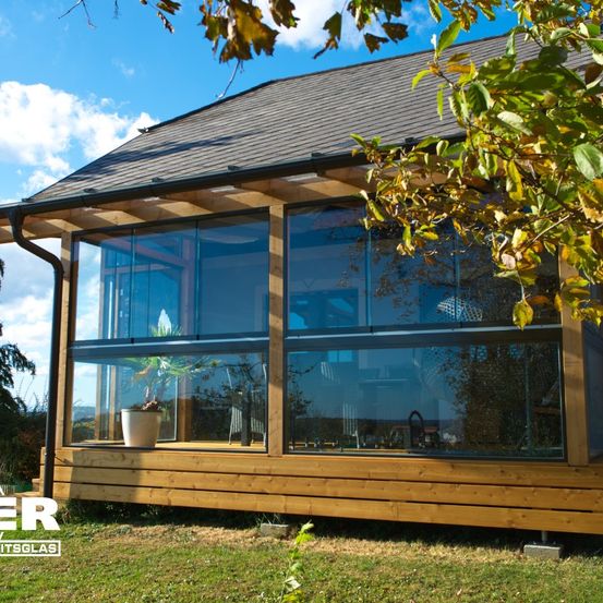Ein modernes Holzhaus mit großen Glasfenstern, umgeben von Grün. Die Fenster reflektieren die umliegende Landschaft, einschließlich eines Baumes mit Herbstblättern.