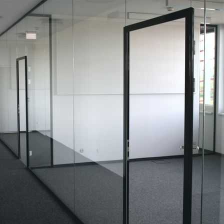 Ein leeres Büro mit Glaswänden und schwarzen Rahmen, ausgestattet mit einem grauen Teppich und einer geschlossenen Tür auf der linken Seite.