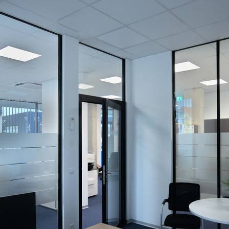 Ein Büroinnenraum mit Glaswänden und Trennwänden, ausgestattet mit einem schwarzen Stuhl, einem weißen Schreibtisch und einem Topf mit Pflanze. Der Raum ist gut beleuchtet mit Deckenleuchten.