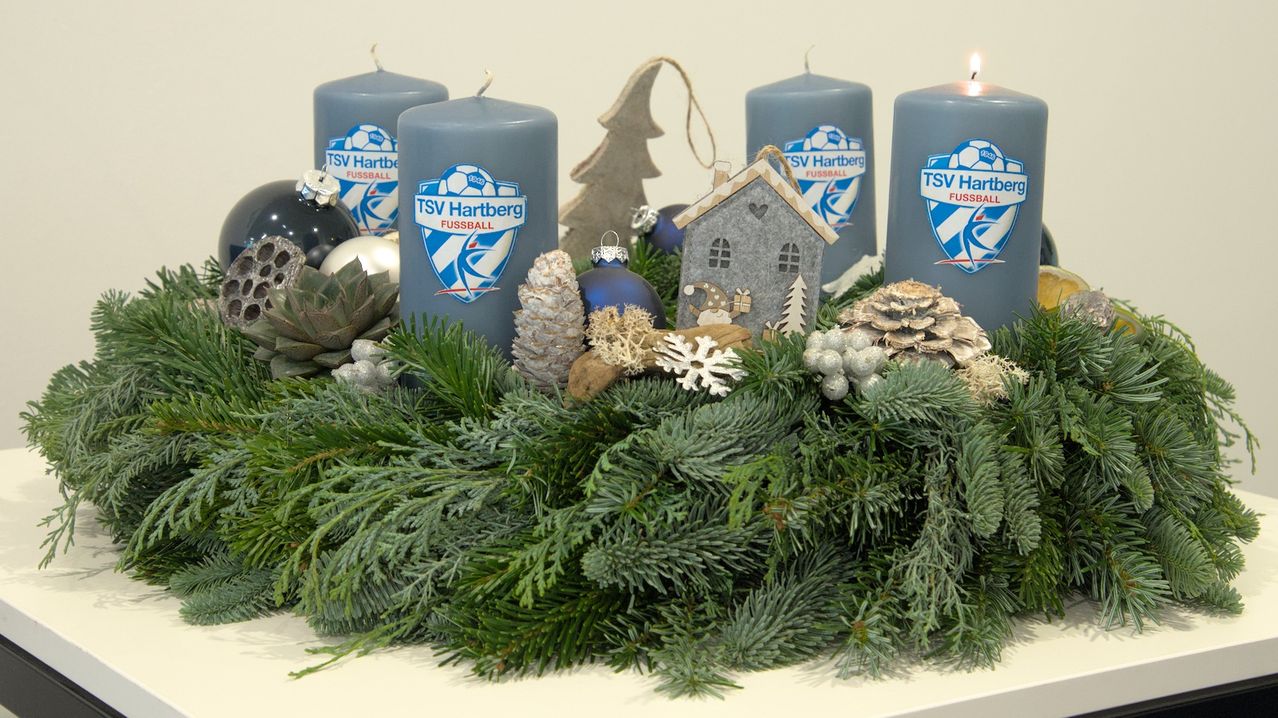 Ein Kranz mit grünen Kiefernzweigen, vier blauen Kerzen, Tannenzapfen, Schneeflocken und einem kleinen Haus steht auf einem weißen Tisch. Die Kerzen tragen das TSV Hartberg Fußballlogo.