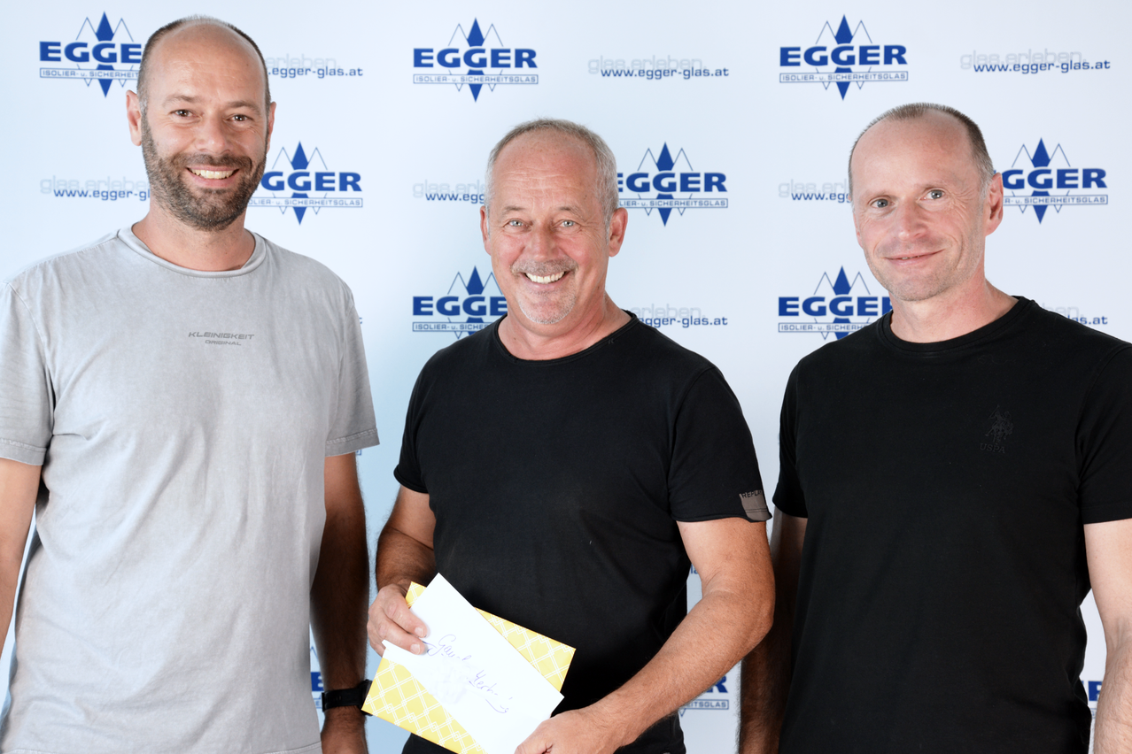 Three men in front of a white backdrop with a blue logo. The middle man holds a yellow envelope, and all three are smiling.