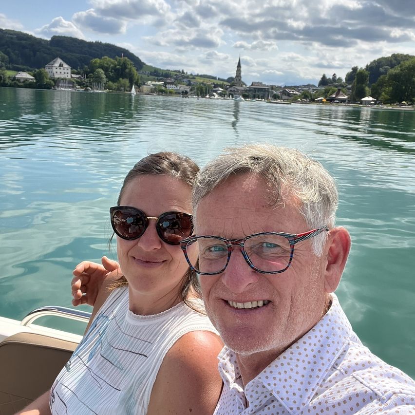 Ein Mann und eine Frau lächeln und posieren für ein Foto auf einem Boot auf einem See mit Bergen im Hintergrund.
