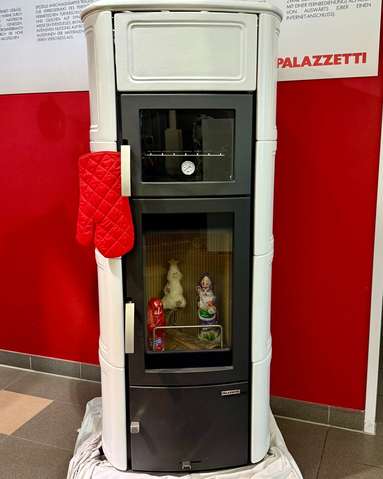 Ein moderner Pailazzi-Ofen mit Backofen und Glastür. Im Inneren befinden sich ein kleiner Weihnachtsbaum und zwei dekorative Figuren. Ein Ofenhandschuh hängt am Griff.