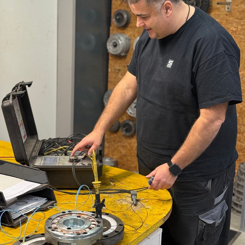 Ein Mann in einem schwarzen T-Shirt arbeitet in einer Werkstatt an einer Maschine, hält ein Werkzeug und untersucht ein Gerät auf einem gelben Tisch.