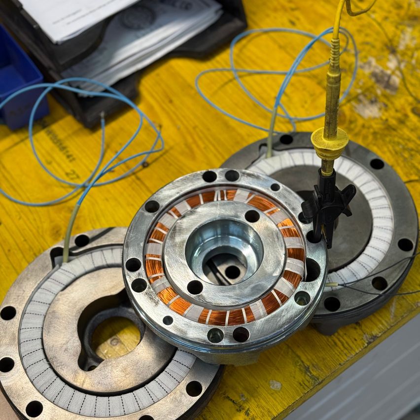 Ein zerlegter Motor mit mehreren Teilen auf einer gelben Arbeitsfläche, angeschlossenen Kabeln und einem Schraubendreher in der Nähe.