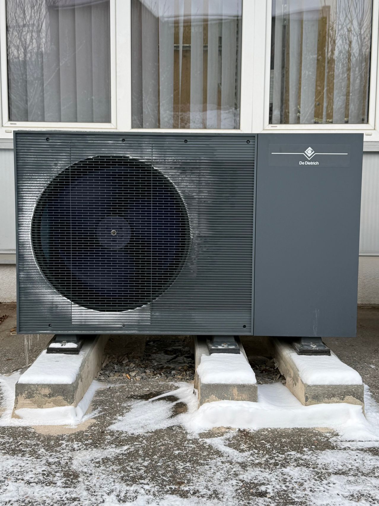 Eine schwarze Außenheizungseinheit steht auf Betonblöcken, mit Schnee bedeckt, hinter ihr befindet sich ein Fenster mit Jalousien.