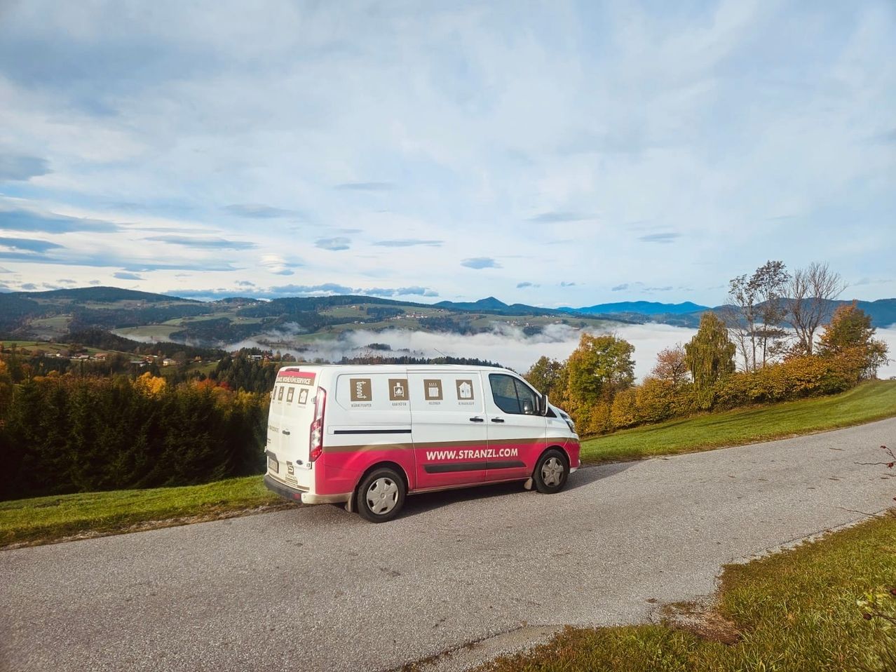 Ein weißer und rosafarbener Van mit dem Text WWW.STRANZL.COM ist auf einer Straße in einem hügeligen Gebiet mit Nebel geparkt.