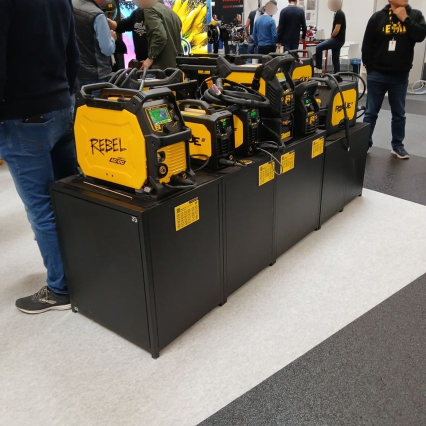 Mehrere gelbe und schwarze Generatoren mit der Aufschrift REBEL sind auf einem schwarzen Ständer ausgestellt, um die sich Menschen versammelt haben.