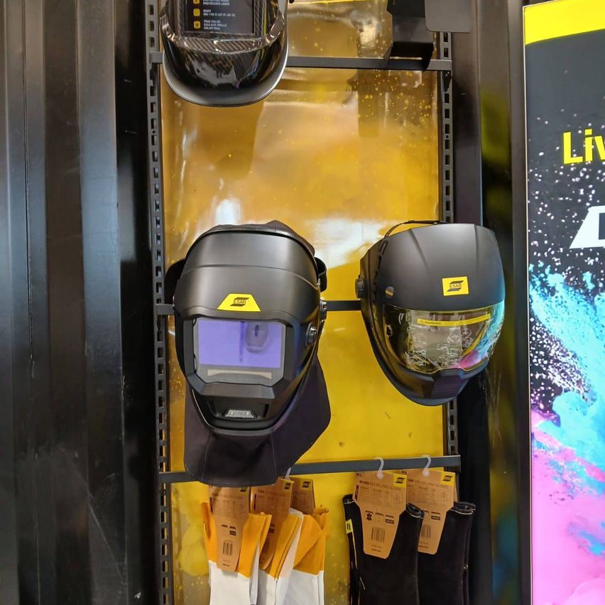 Drei Schweißhelme sind auf einem gelben Regal ausgestellt. Der oberste Helm ist schwarz mit einem gelben Logo. Darunter hängen zwei weitere schwarze Helme mit gelbem Logo.