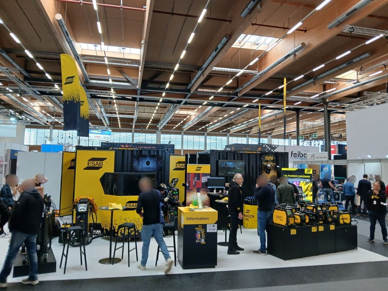 Eine Messe mit gelben Ständen und Bannern, darunter ein Mann, der sich in der Nähe eines Tisches mit einem gelben Schild befindet. Andere Menschen sind um eine Ausstellung von Maschinen versammelt.