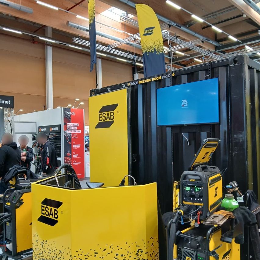 Ein Innenbereich mit einem gelben Stand und einem schwarzen Stand, auf dem die Marke ESAB angezeigt wird. Ein Monitor befindet sich auf dem schwarzen Stand, und einige Schweißgeräte stehen auf dem Boden.