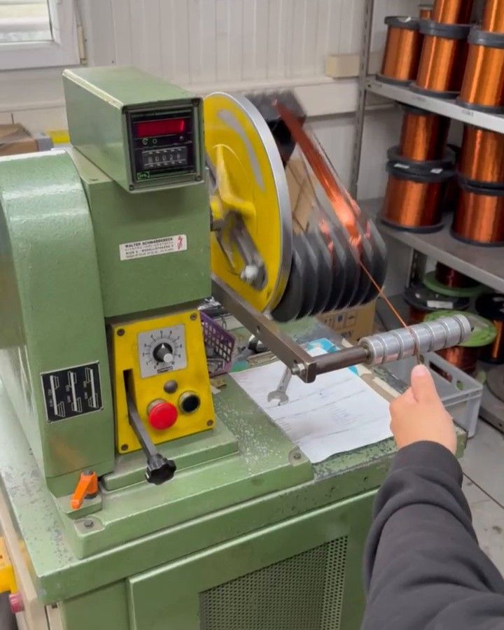 Eine Person bedient eine grüne Maschine mit einem digitalen Display, drückt einen roten Knopf und managt Kupferdrähte in einer Fabrikhalle.