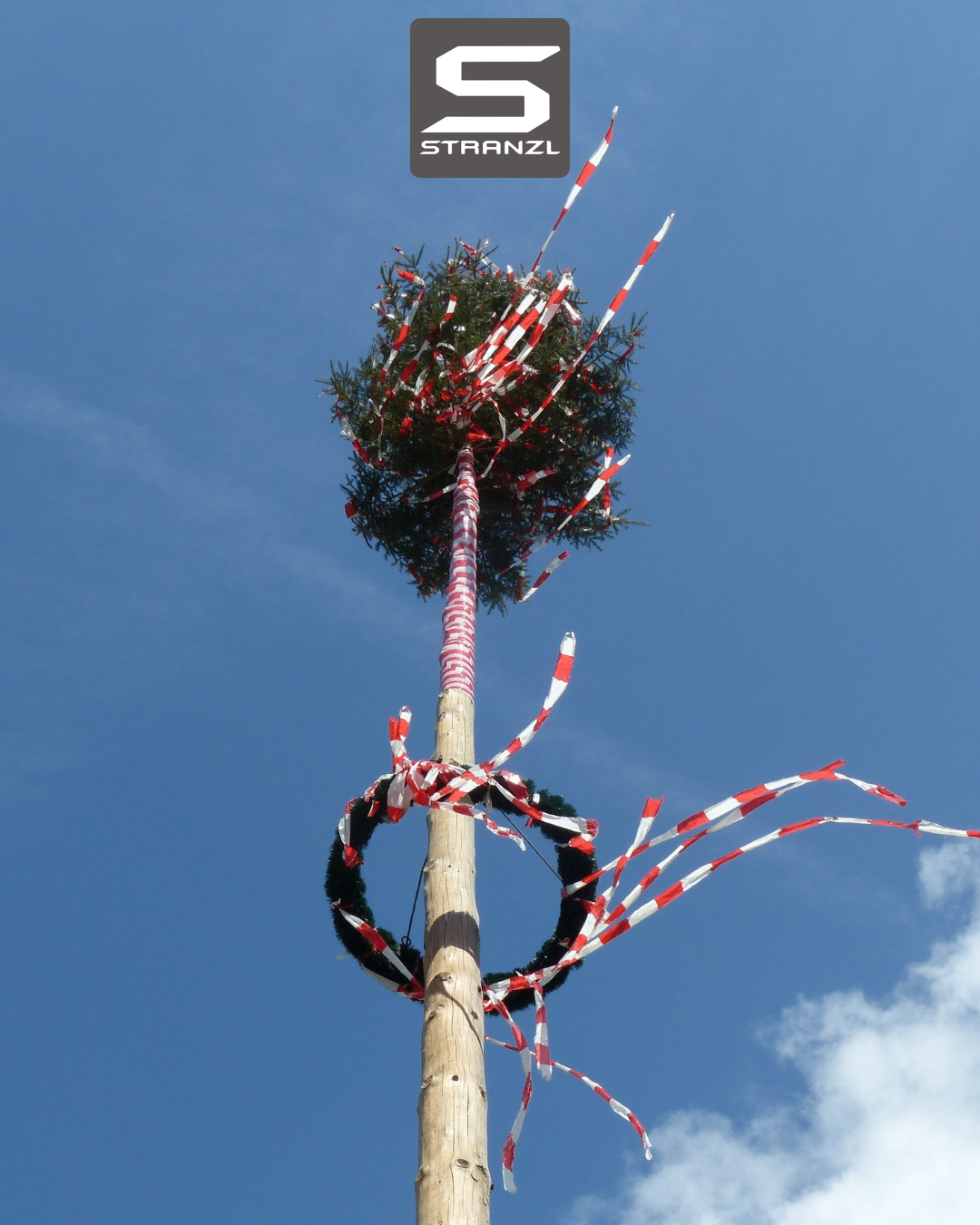 Ein hoher Holzpfahl mit einem mit roten und weißen Bändern geschmückten Tannenbaum. Der Pfahl ist mit Bändern und zwei Kränzen geschmückt. Der Himmel ist blau.