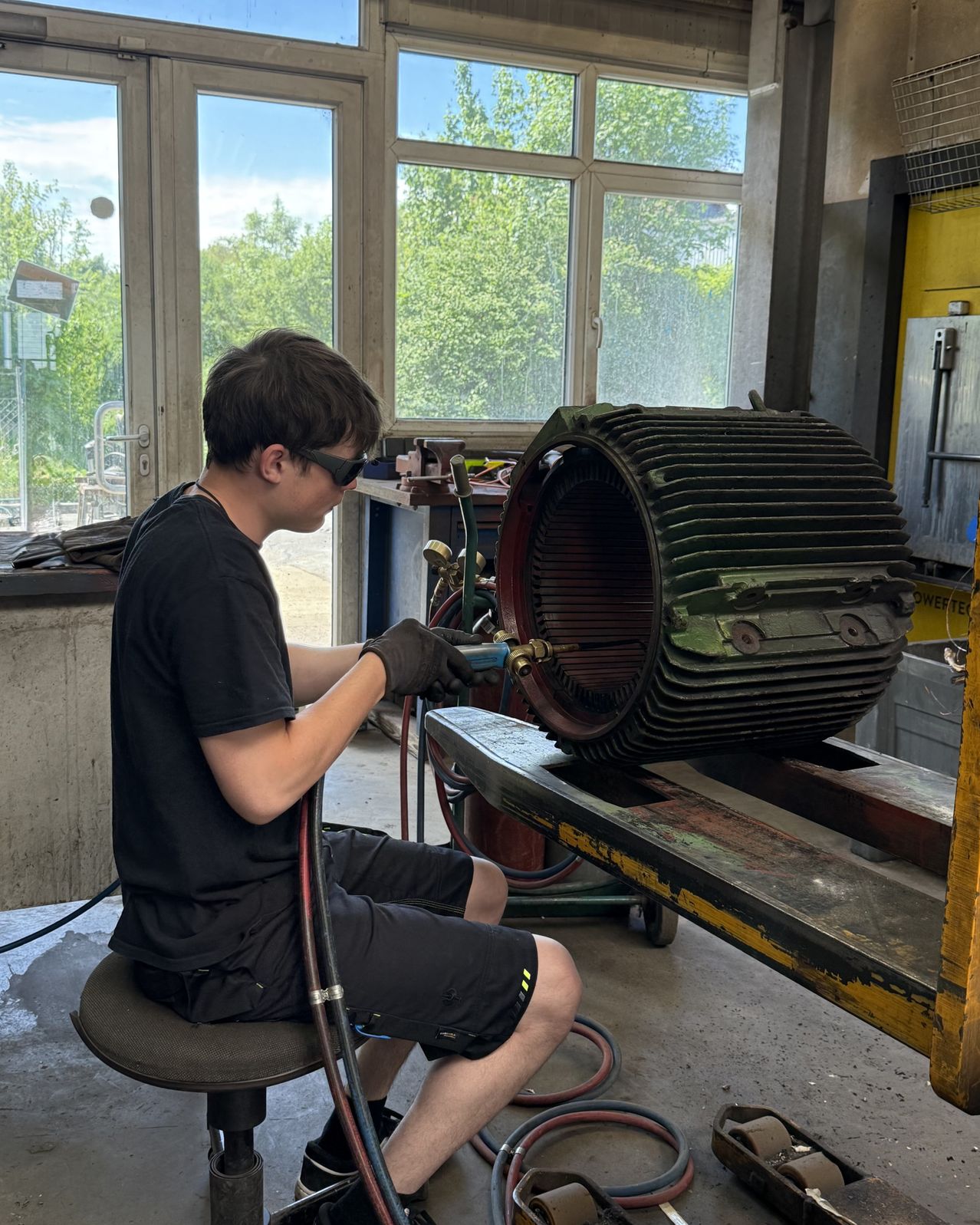 A young man wearing a black shirt, shorts, and sunglasses works on a large, complex machine in a workshop.