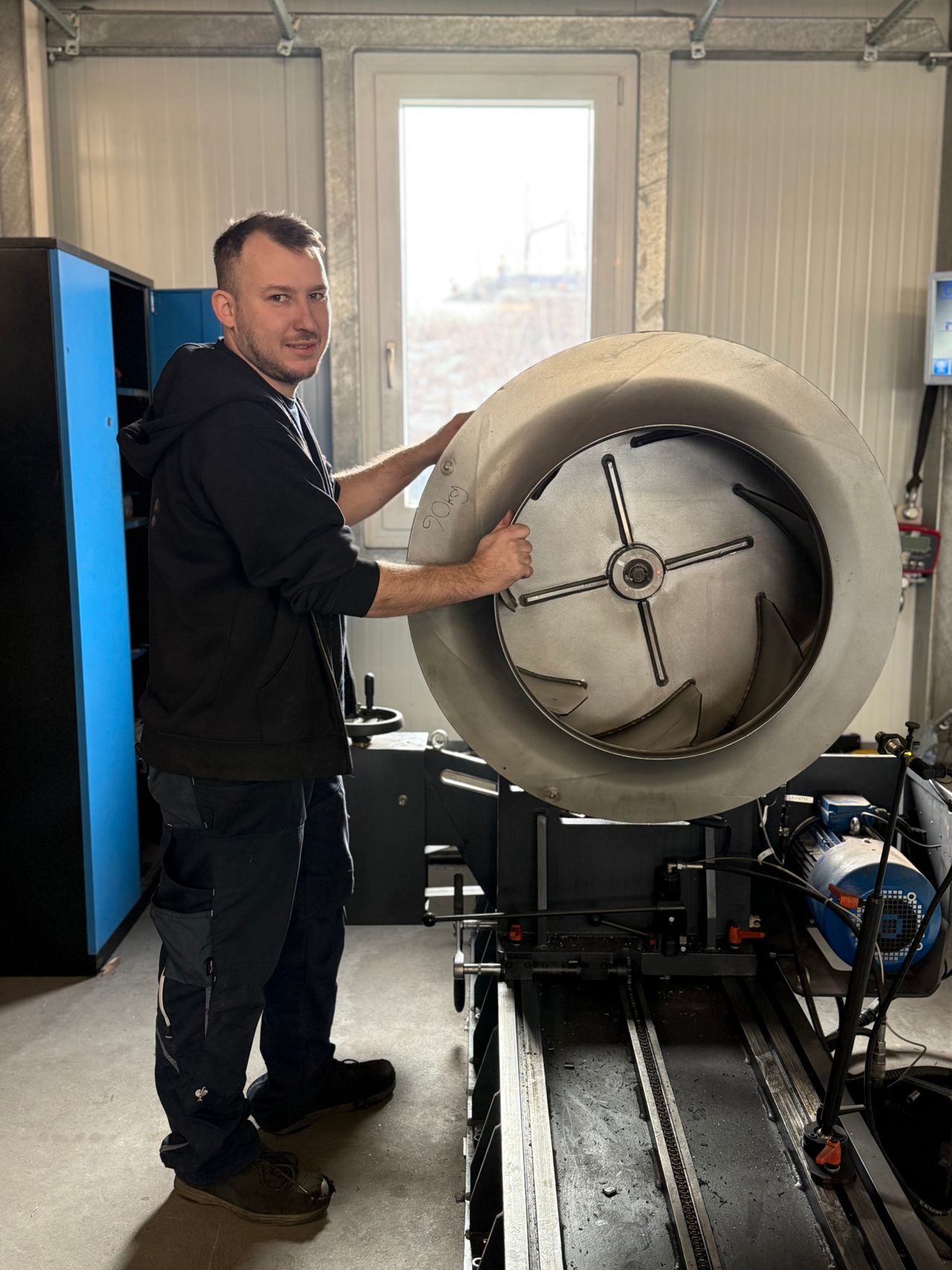 A man in a black jacket and pants holds a large metal object with his hands. He is in a workshop with a window in the background.