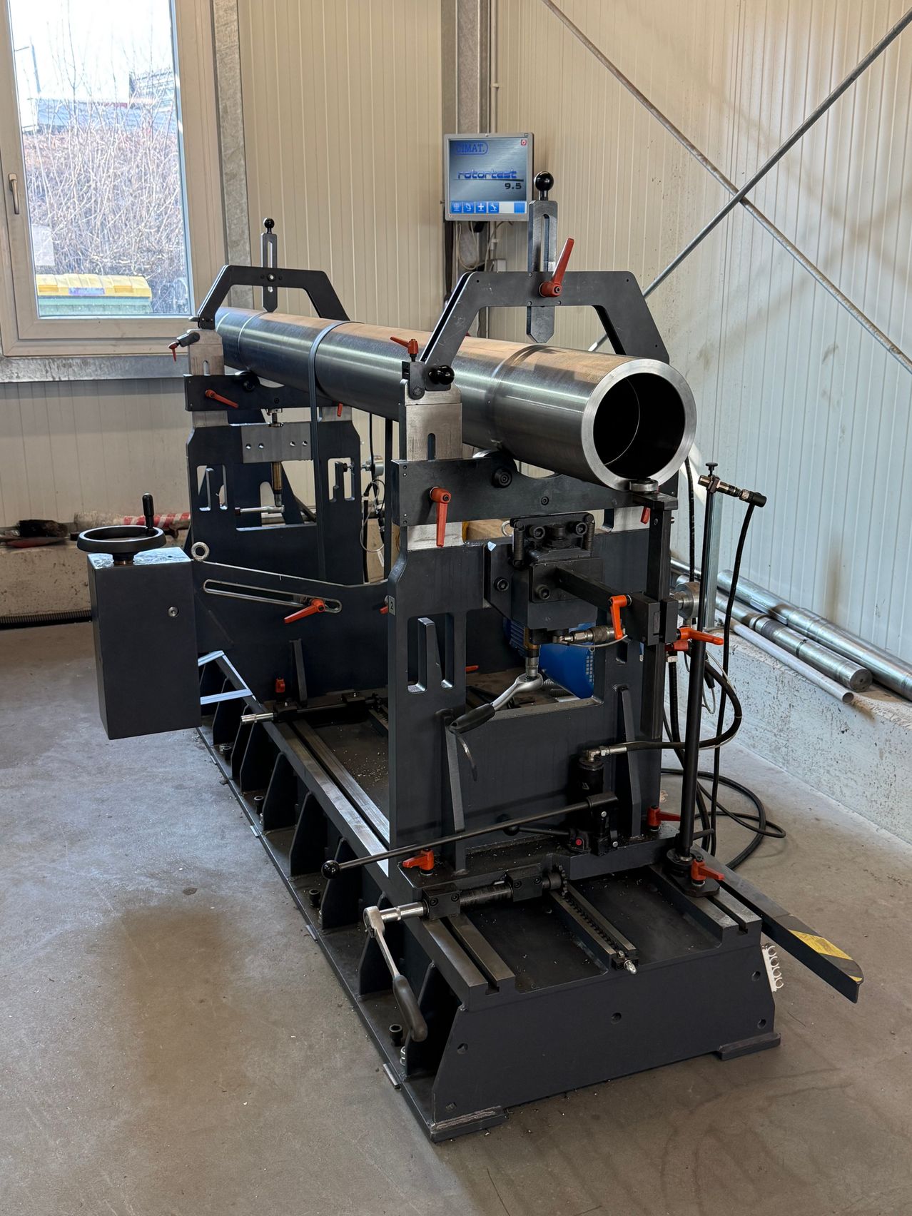 A large metal tube is being processed by a machine in a workshop. The machine has a digital display, various tools, and a window showing a view outside.