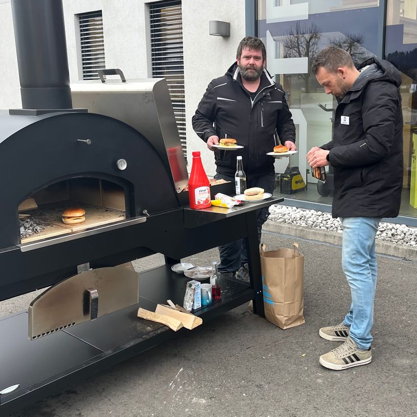 Zwei Männer stehen bei einem großen schwarzen Pizzaofen. Einer hält einen Teller mit Burgern, der andere eine Flasche. Ein brauner Papierbeutel ist in der Nähe.