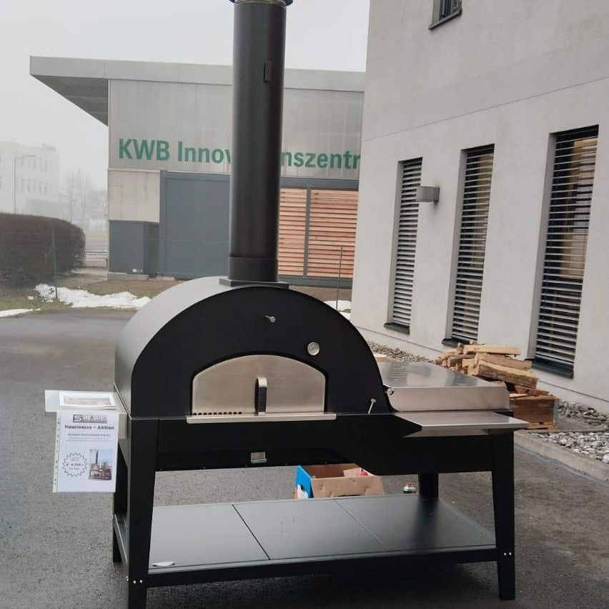 Ein schwarzer Outdoor-Pizzaofen steht vor einem Gebäude. Ein Schild mit Text ist am Ofen befestigt. In der Nähe befinden sich einige Holzkisten und ein Gebäude mit der Aufschrift 'KWB Innov'.