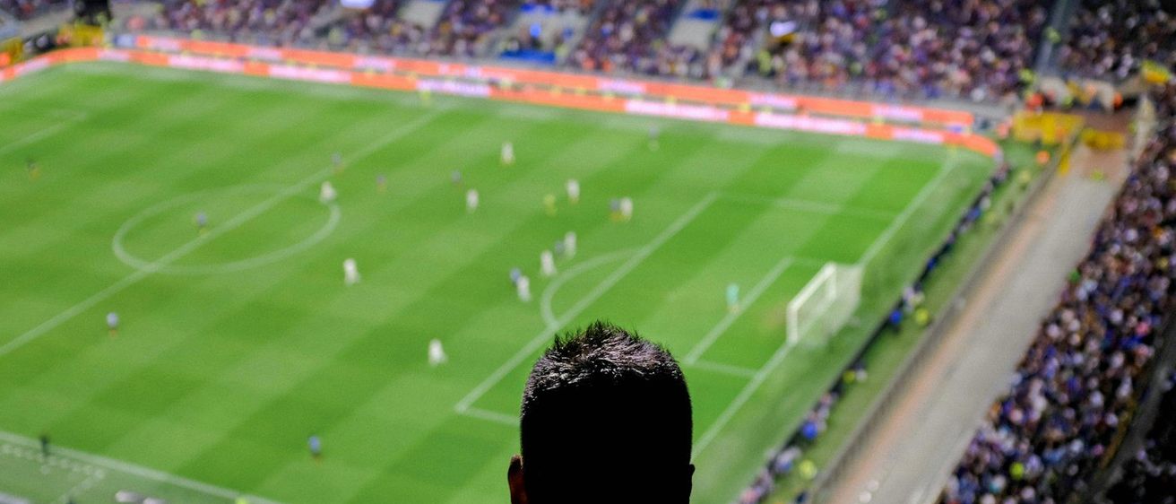 Ein volles Stadion mit vielen Zuschauern, die bei Nacht ein Fußballspiel verfolgen. Die Spieler sind auf dem Feld, und die Atmosphäre ist lebhaft.