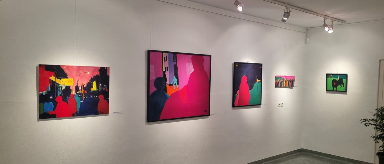An einer weißen Wand sind mehrere Gemälde mit leuchtenden Farben zu sehen. Das größte Gemälde zeigt eine rosafarbene Figur vor einem Stadthintergrund. Links davon befindet sich ein kleineres Gemälde mit einer roten Figur. Rechts hängt ein Gemälde mit einer roten Figur. Oben hängt eine kleine Gruppe bunter Gemälde.