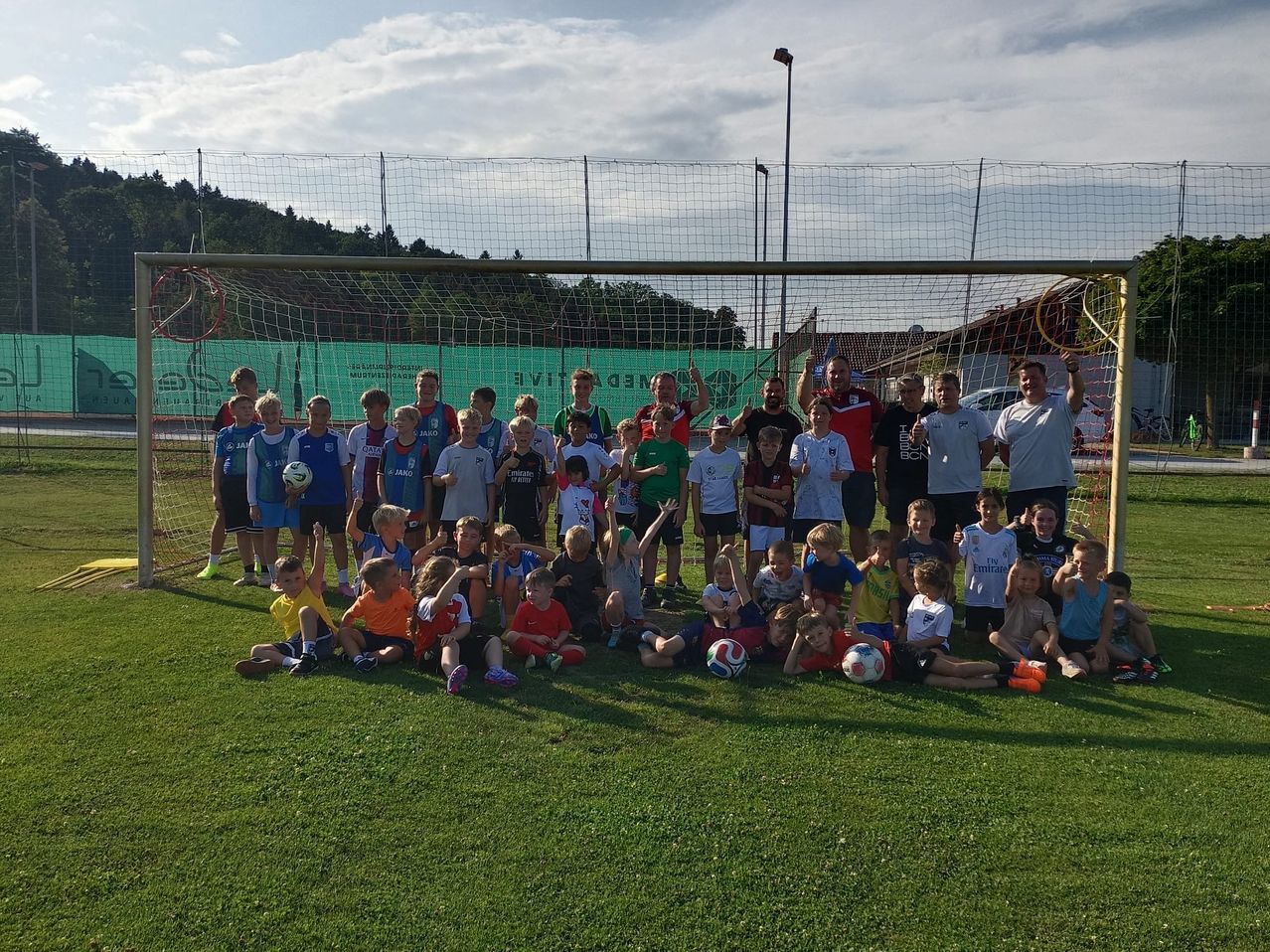 Eine Gruppe von Kindern und Erwachsenen posiert für ein Foto vor einem Fußballtor, wobei einige Fußballbälle halten und andere ihre Hände heben.