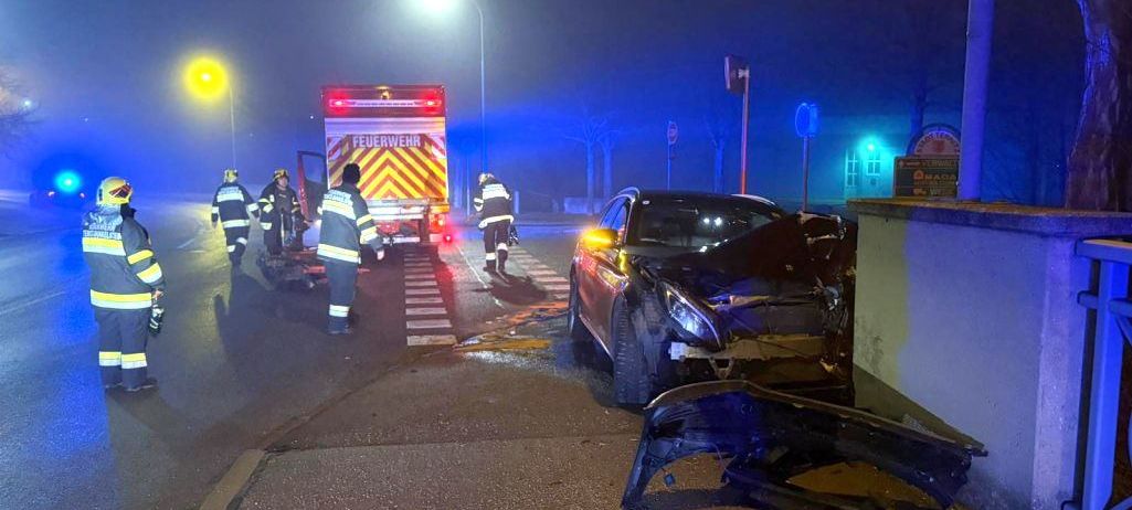 Einsatzkräfte bei Nacht, die sich um ein demoliertes Auto mit abgerissener Frontstoßstange auf einer Straße kümmern, mit einem Feuerwehrwagen dahinter.