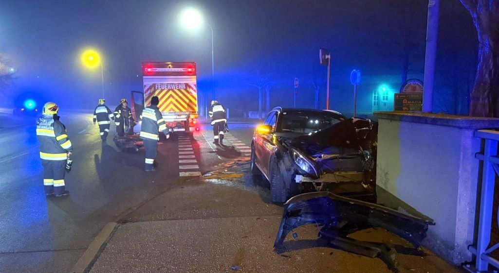 Einsatzkräfte bei Nacht, die sich um ein demoliertes Auto mit abgerissener Frontstoßstange auf einer Straße kümmern, mit einem Feuerwehrwagen dahinter.