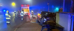 Einsatzkräfte bei Nacht, die sich um ein demoliertes Auto mit abgerissener Frontstoßstange auf einer Straße kümmern, mit einem Feuerwehrwagen dahinter.
